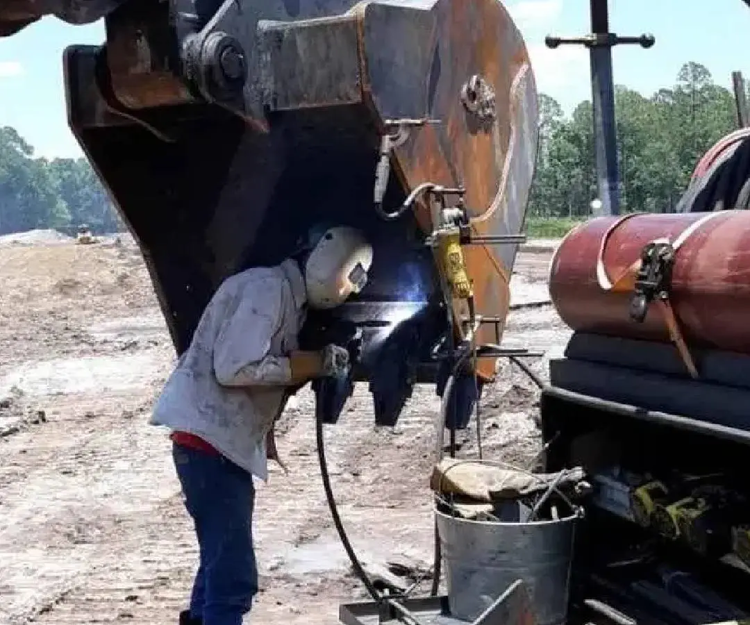 Worker welding metal on heavy machinery outdoors.