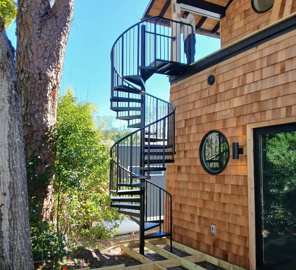Black metal spiral staircase outside a wooden house surrounded by greenery.